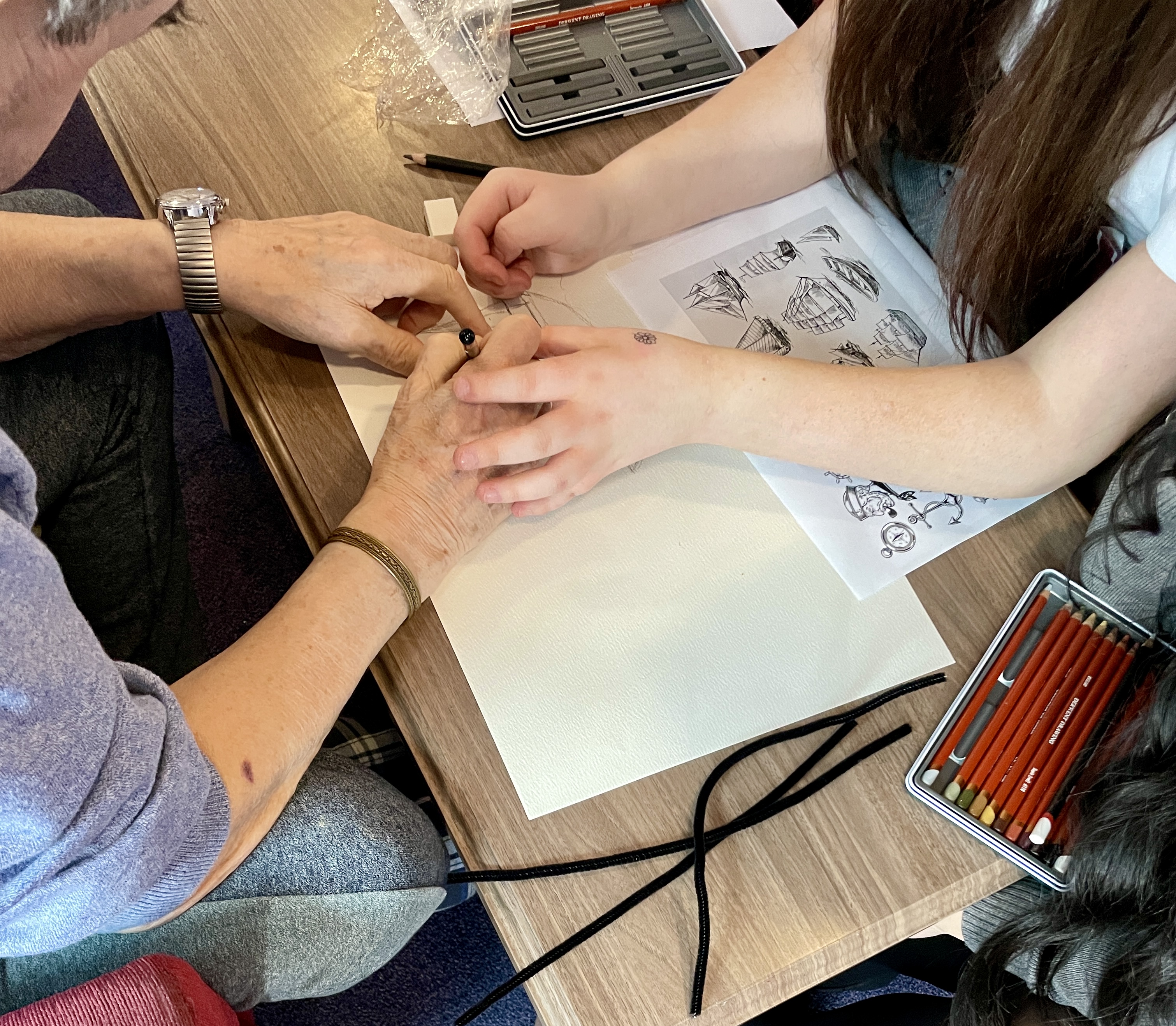 Young person's hands assisting older hands to draw a ship using a pencil and paper