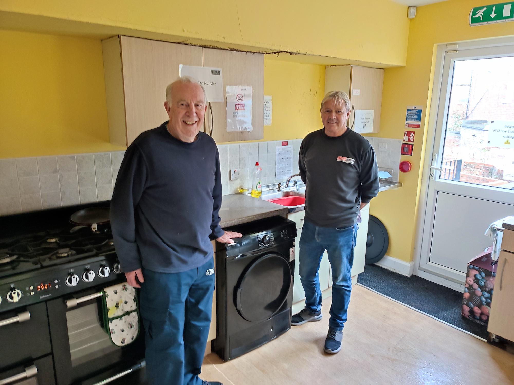 Two men standing in a bright kitchen with a tumble dryer between them