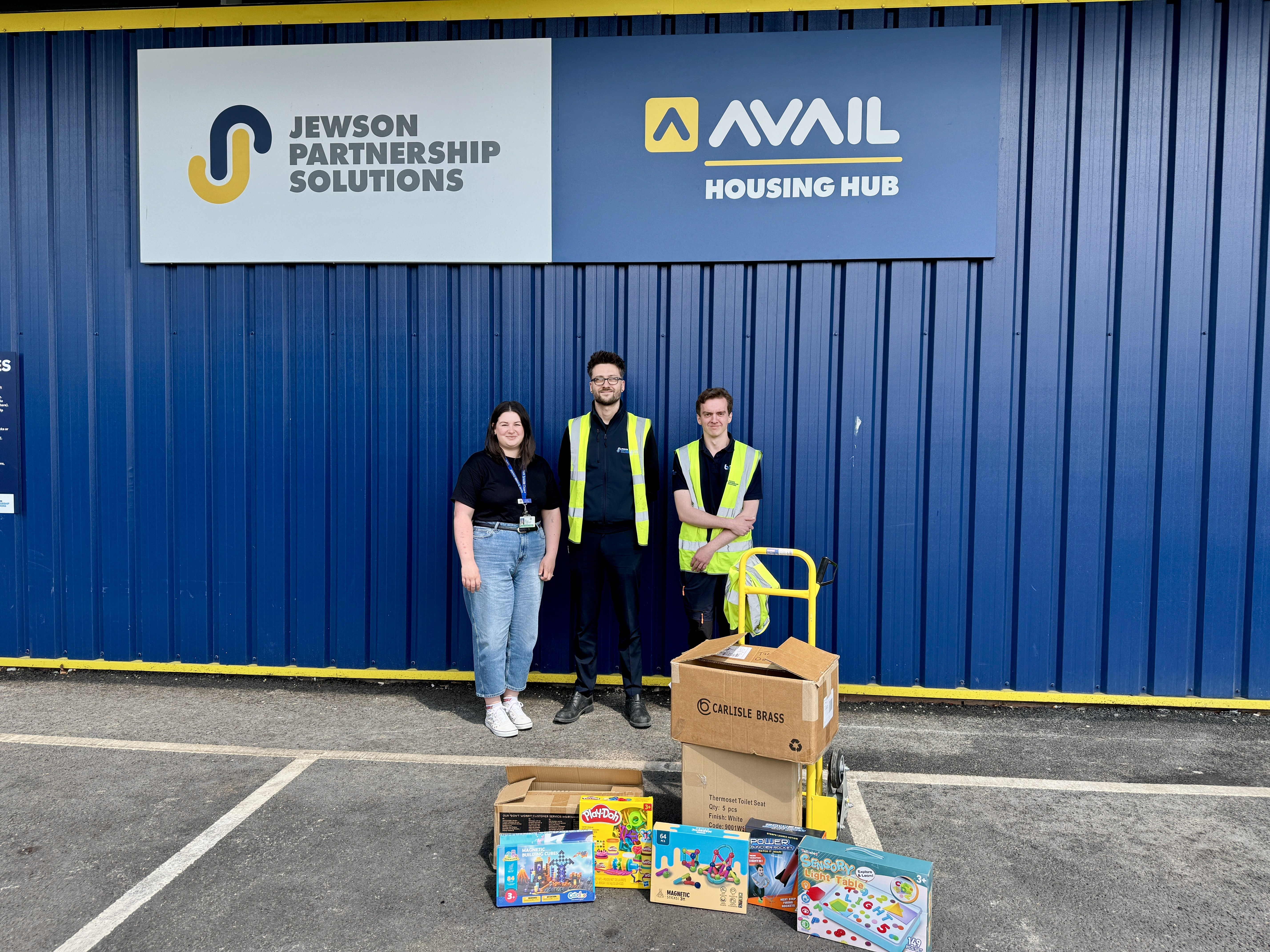 Three people standing behind a pile of gifts with signs behind them reading Jewsons and Avail Housing hub