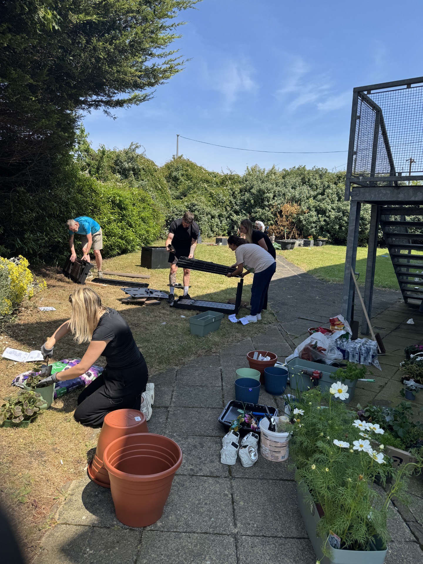 Several people work together in an outdoor garden space on a sunny day, assembling furniture and planting flowers. In the foreground, a person kneels on the grass filling a planter with soil beside large plant pots, while others assemble a black bench or shelving unit on a paved path. Gardening supplies, potted flowers, tools, and containers are arranged around the area. The space is bordered by mature shrubs and trees, with a metal staircase visible on the right, showing a community landscaping and improve