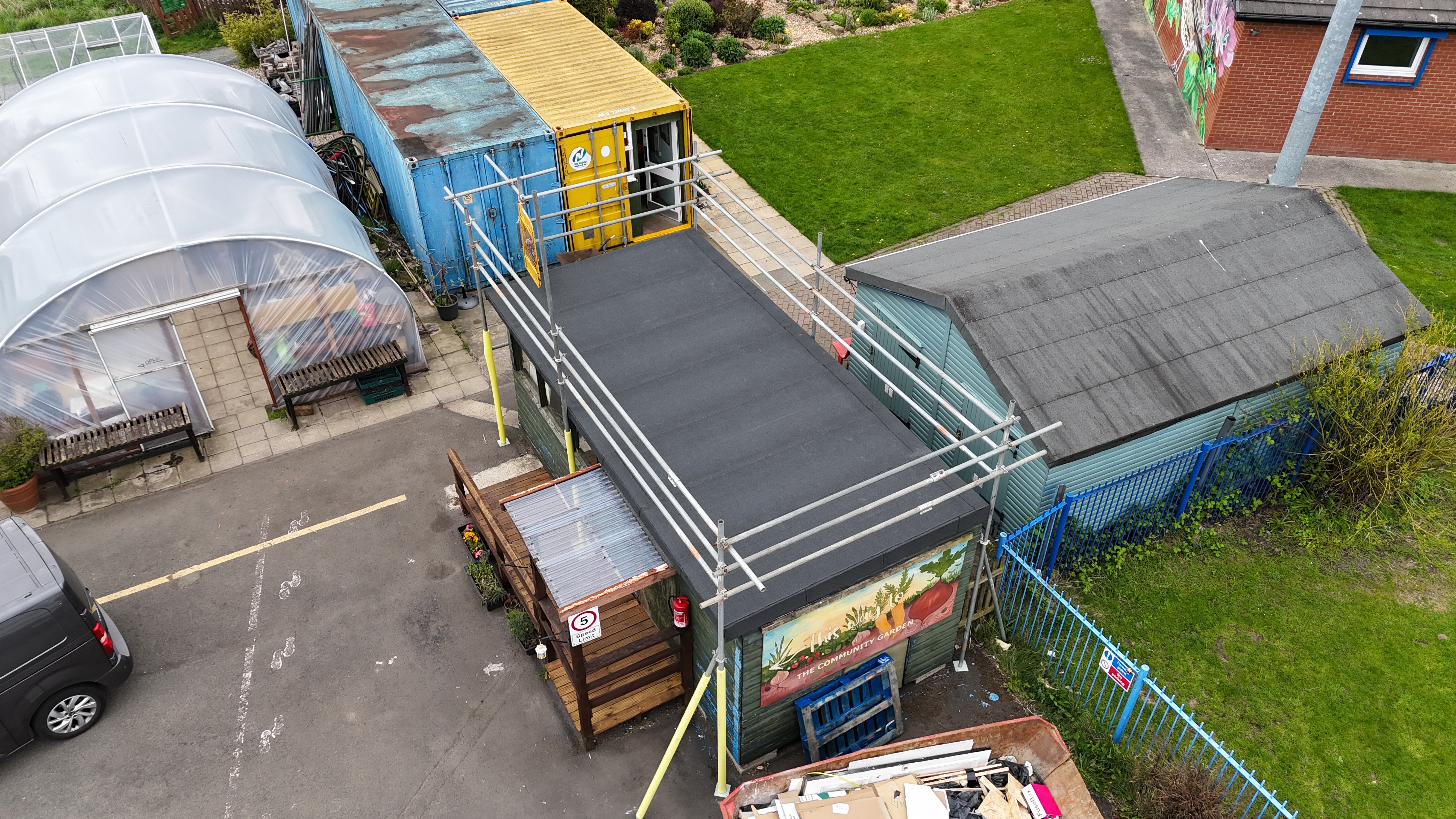 Aerial view of a community garden featuring a polytunnel greenhouse, storage buildings, shipping containers, and a small garden hub building surrounded by scaffolding