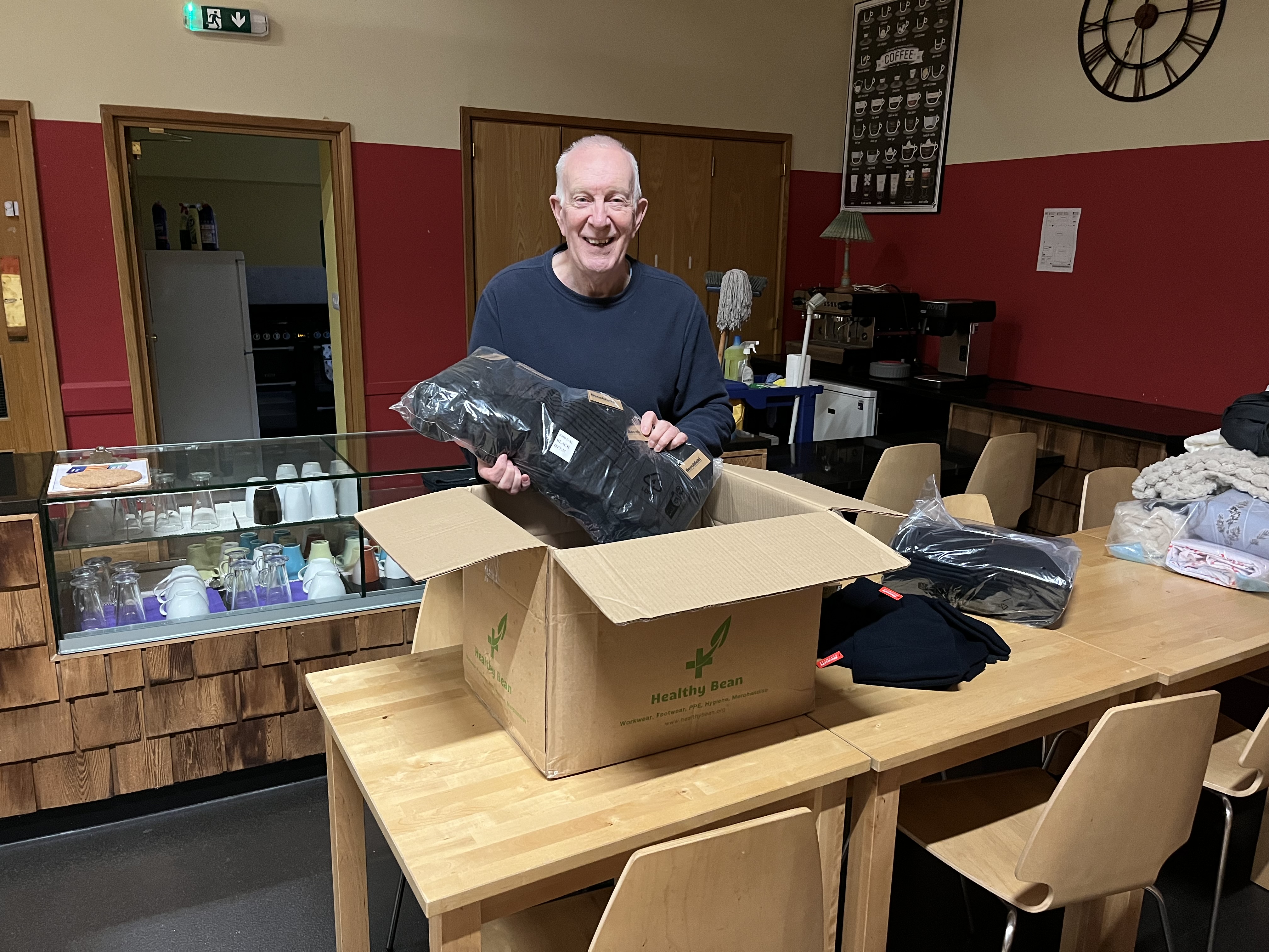 Person unpacking donated clothing from a box in a community café, with additional garments laid out on tables and catering facilities in the background