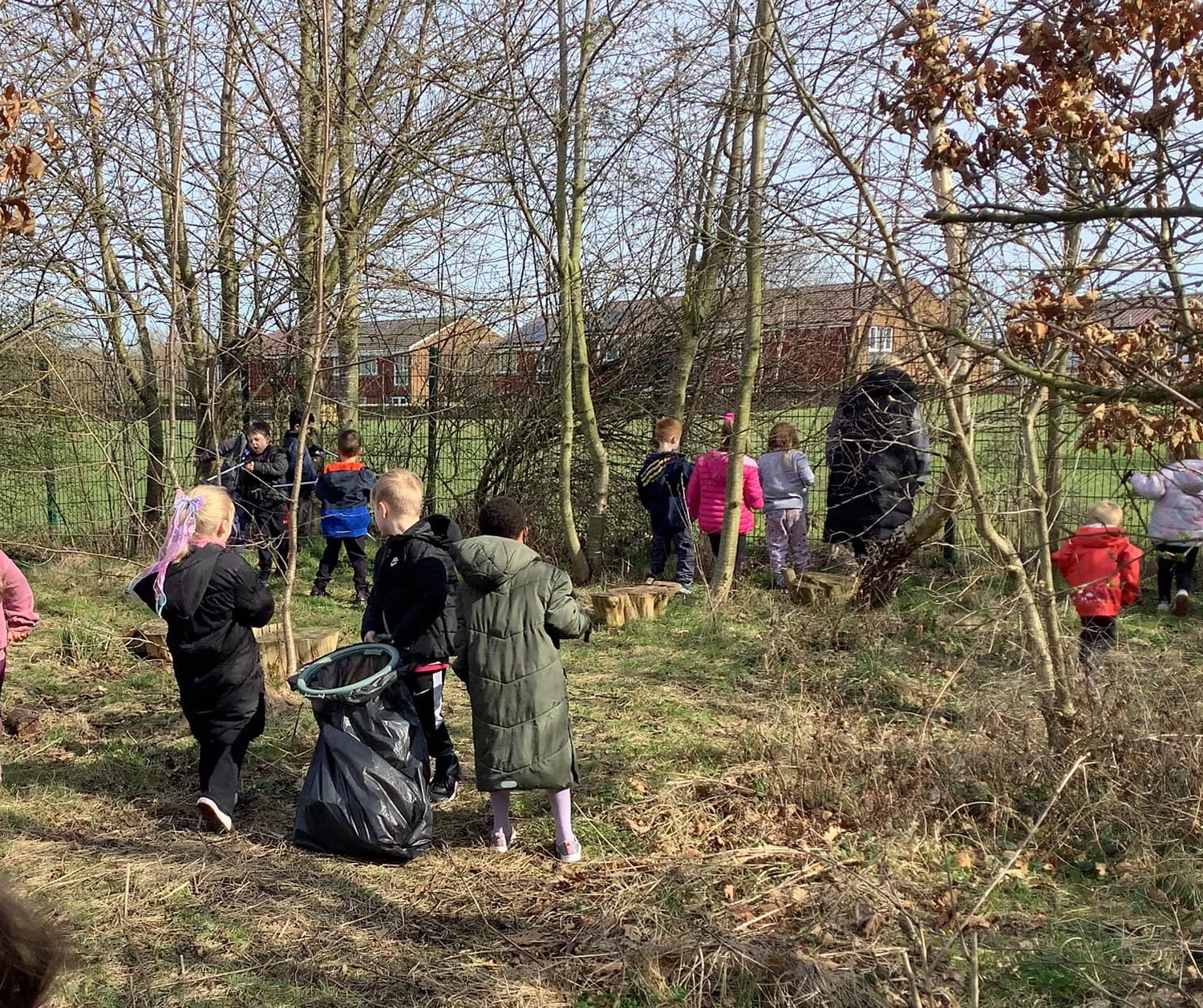 Children and adults participating in a woodland litter-picking and nature activity, carrying rubbish bags through a wooded area next to a grass field