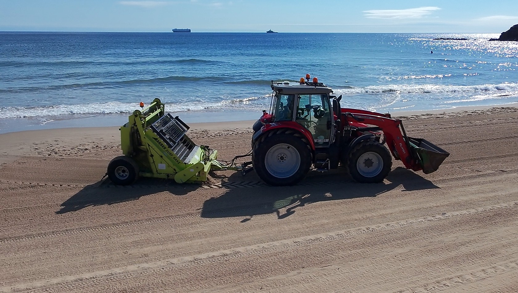 Red tractor on a beach with a green mechanical rake attached to the back. The sea is in the bacground