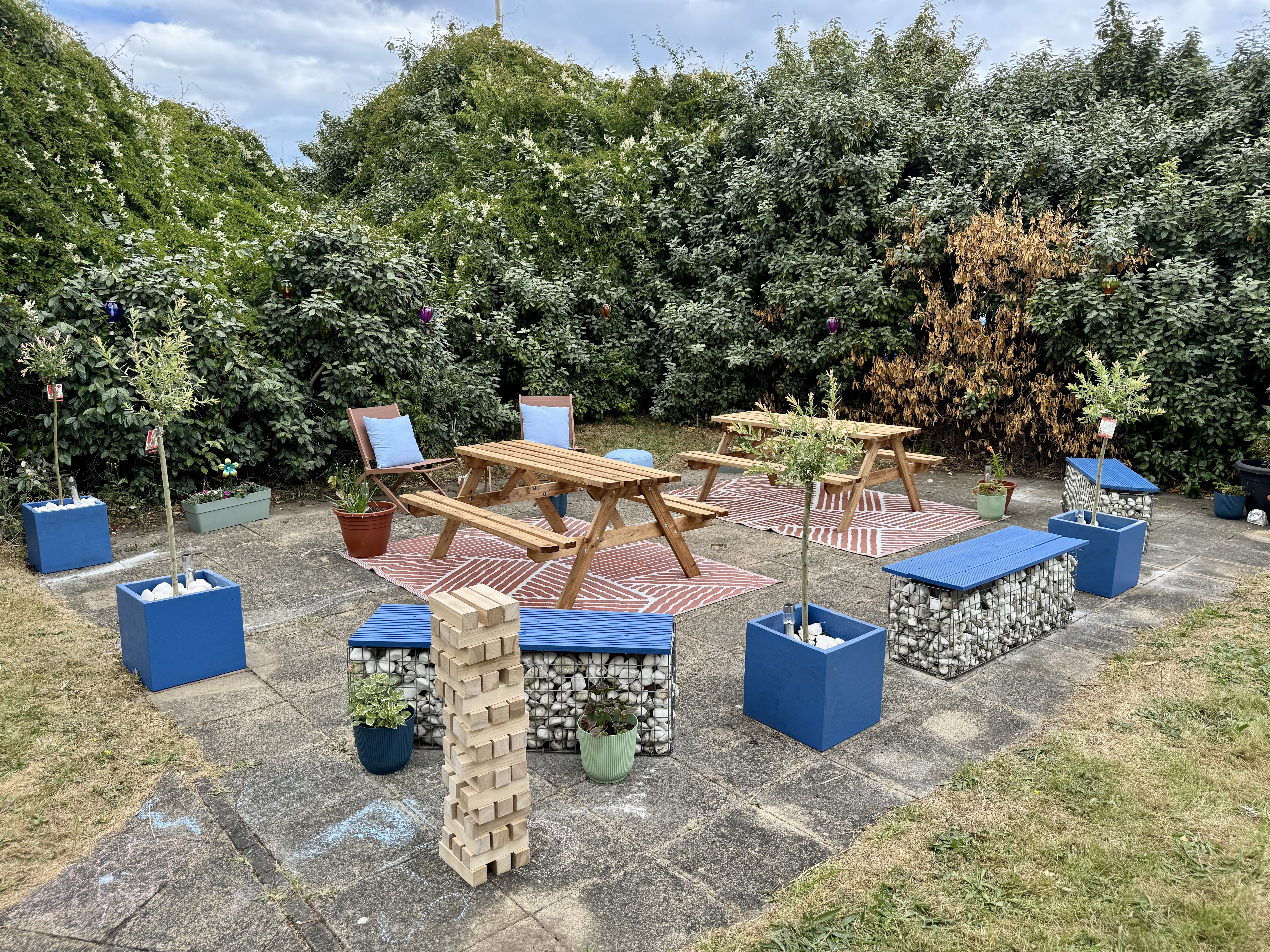 An outdoor community garden seating area on a paved patio, surrounded by dense green shrubs and climbing plants. Two wooden picnic benches sit on patterned outdoor rugs, with additional blue benches, planters containing small trees and flowers, and decorative gabion-style seating filled with stones. A stack of oversized wooden blocks stands in the foreground, and potted plants are placed throughout the space. The area appears designed for social gatherings and outdoor activities in a landscaped garden setti