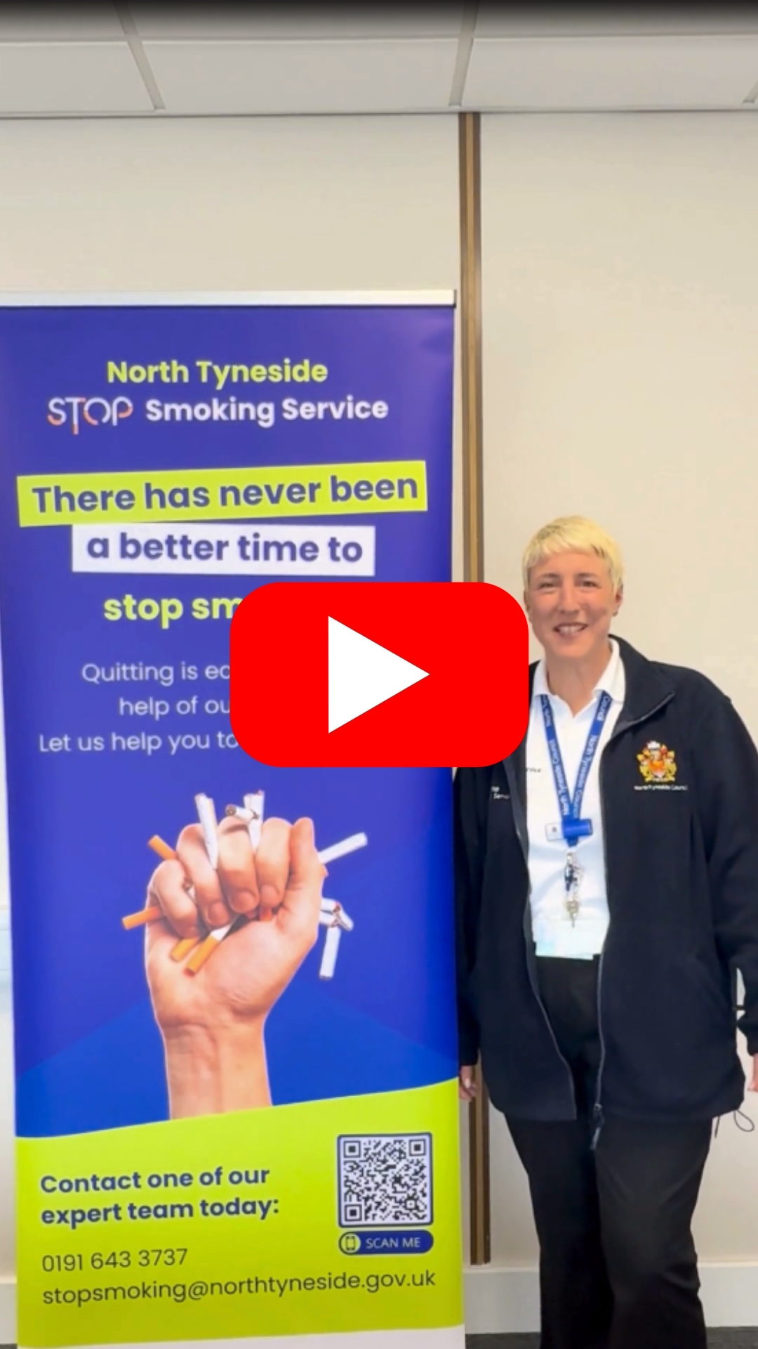 A blonde, short haired woman standing next to a banner which has details on how to contact the North Tyneside Stop Smoking service