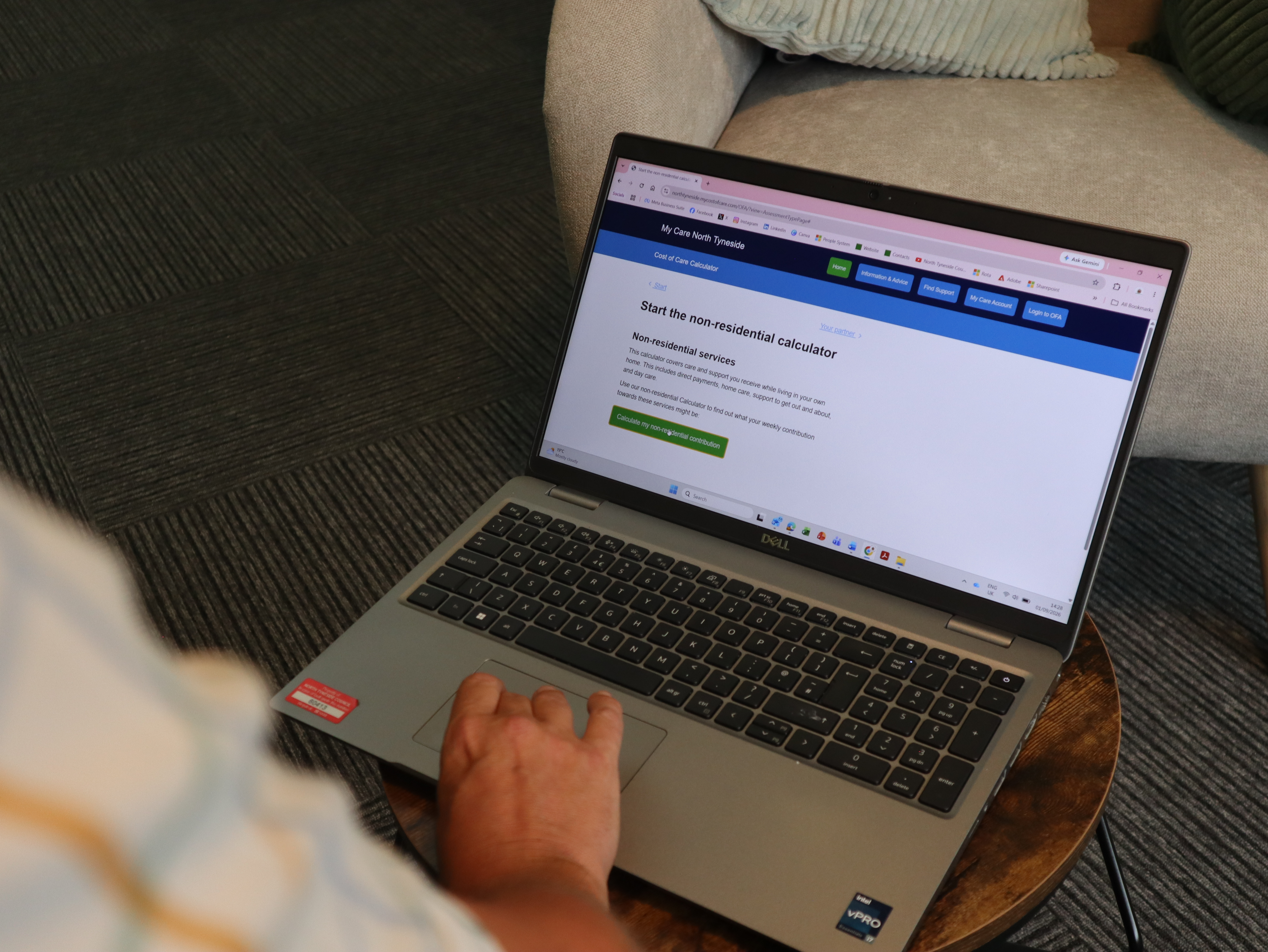 A laptop open showing the cost of care calculator page with a hand operating the mouse, in a room with a comfy chair and coffee table