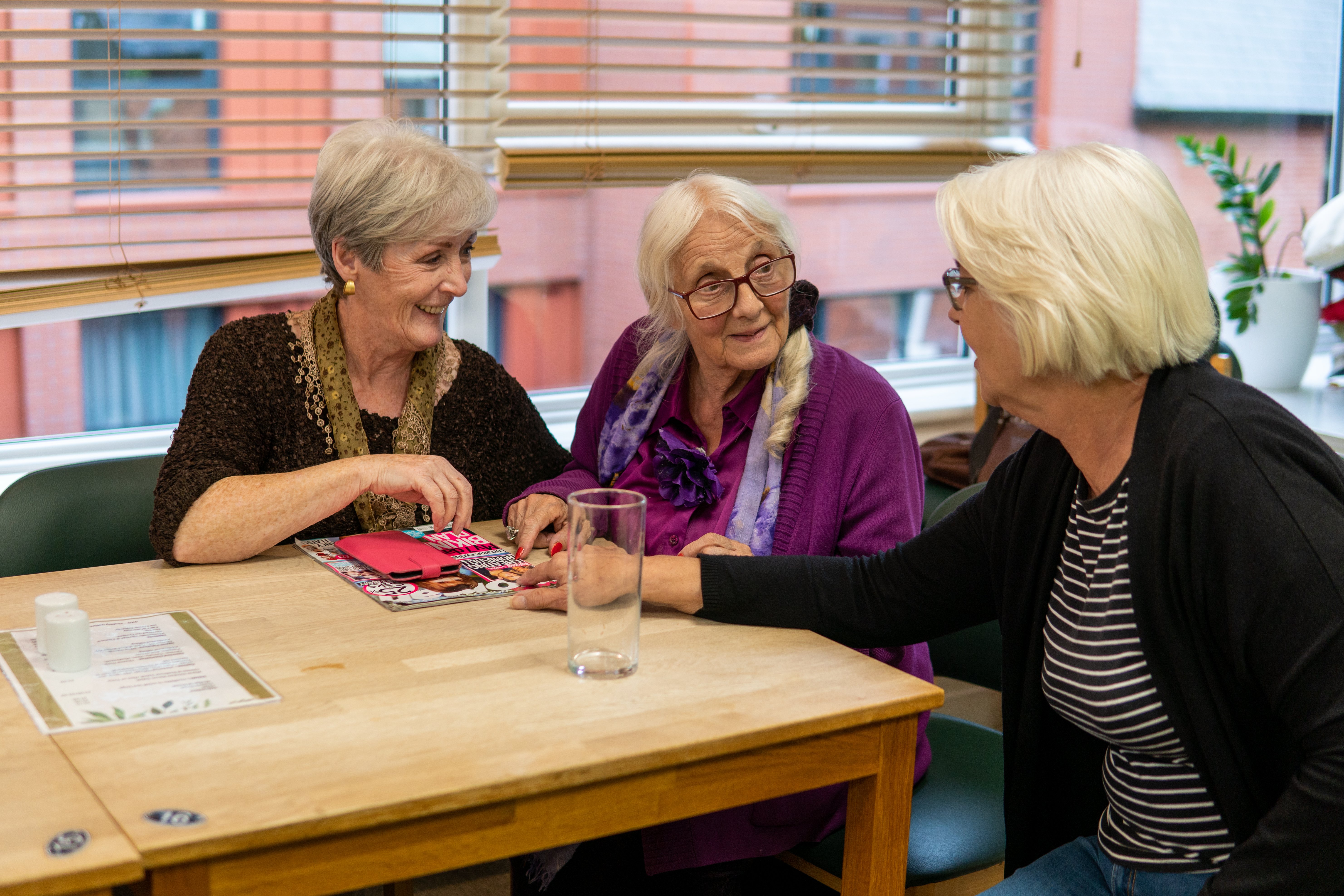 Three older women sitting at a table in a cafe chatting and smiling with a window behind and a view of buildings