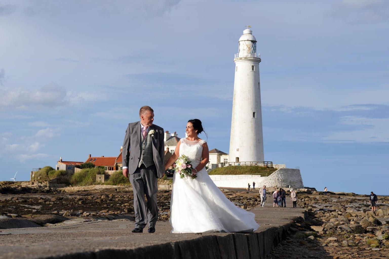 Bride and groom walking away fromt he white lighthouse along a causeway
