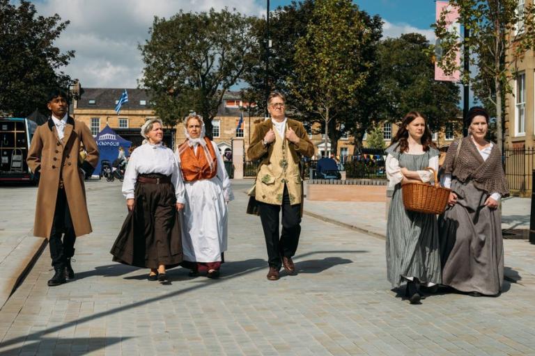 A performance of A Georgian Cluedo with six men and women in period dress walking side by side down Howard Street in the sunshine with the trees of Northumberland Square in the background