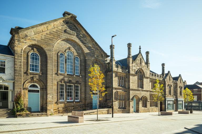 The Exchange Theatre a large historic sandstone building with arched windows with trees and the new pale grey paving on Howard Street in front and a blue sky behind