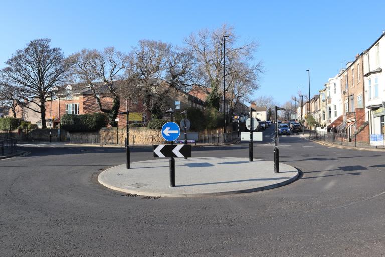 The roundabout at Gateway 3 with keep left signs and a paved centre, there are no cars, with trees behind and a blue sky