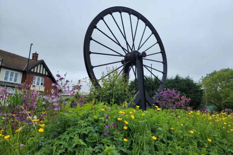 An image of the Burradon Wheel public artwork a big iron pit wheel set in a flowerbed with rooftops visible behind