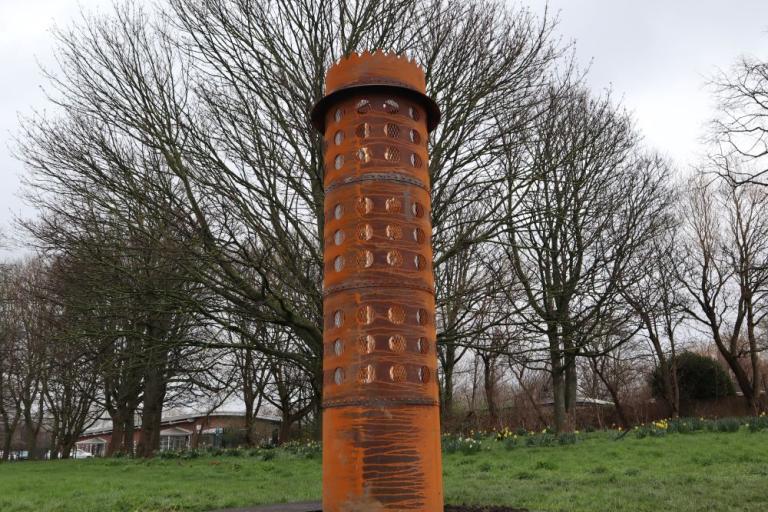 A weather steel tower sculpture in the shape of Stephenson's pit lamp standing on a grassed area with trees behind 
