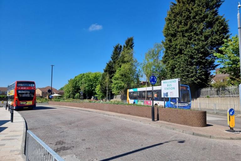 A colour photo of Wallsend bus station with two buses passing each other, blue skies and trees in the background