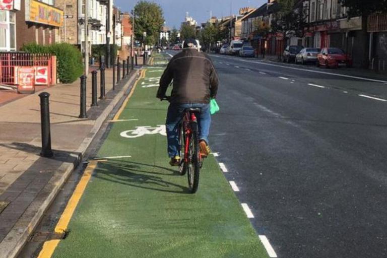 A colour image of a man riding a bike on a green cycle lane on an urban street with shops and houses 