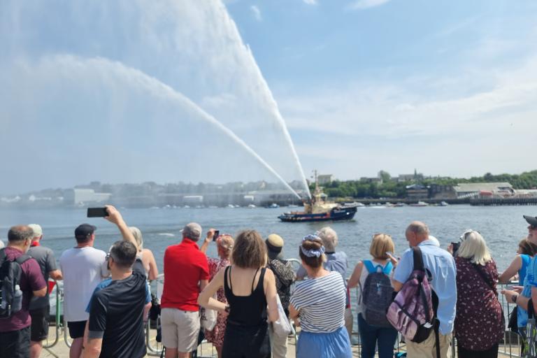 A crowd of people standing on the edge of the Fish Quay watching boats on the river and taking photos as the tug boat sprays water high into the air