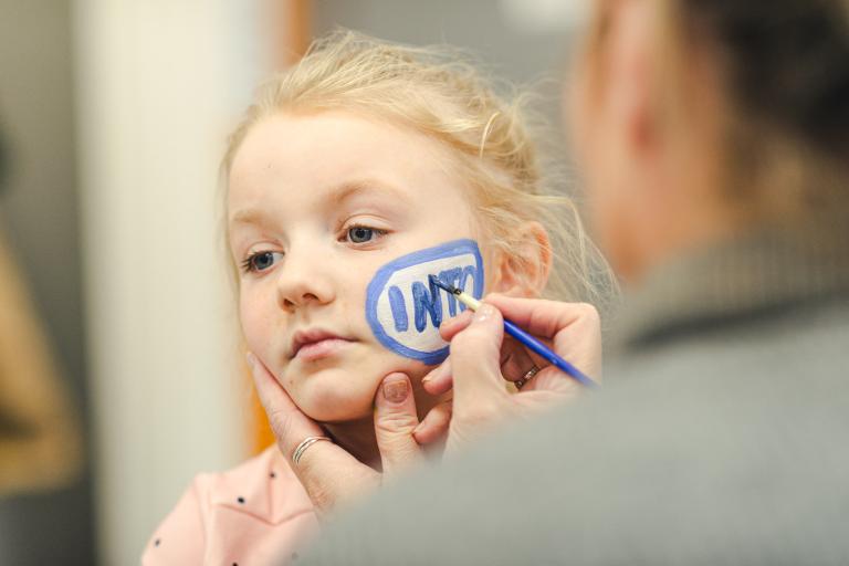 Young blonde girl having her face painted with the easeINTO logo of blue and white