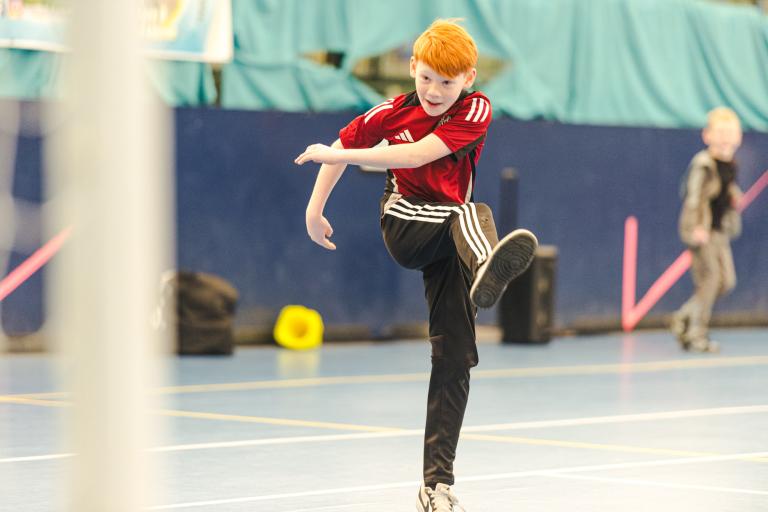 Young person kicking a ball. They're wearing black tracksuit bottoms and a red t shirt