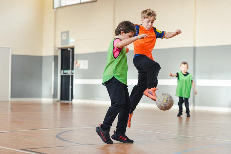 Boys wearing orange and green bibs playing football in a sports hall