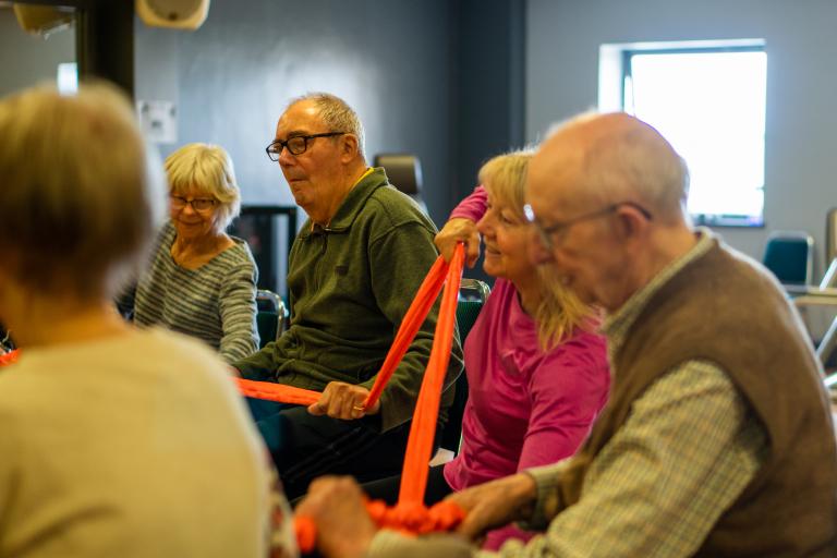 A group of older people taking part in a seated exercise class using a resistance band, three women and two men