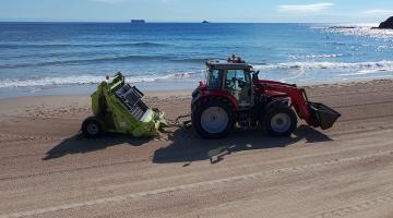 Red tractor on a beach with a green mechanical rake attached to the back. The sea is in the bacground