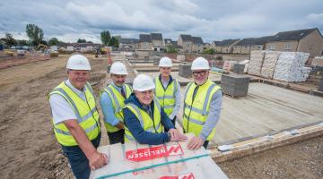 A groupd of five people stand wearing PPE infront of a building site