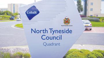 A sign at the entrance to the North Tyneside Council Quadrant offices with the name of the council and surrounded by low shrubs with a road and office buildings in the background