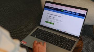 A laptop open showing the cost of care calculator page with a hand operating the mouse, in a room with a comfy chair and coffee table