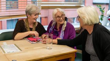 Three older women sitting at a table in a cafe chatting and smiling with a window behind and a view of buildings