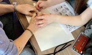 Young person's hands assisting older hands to draw a ship using a pencil and paper