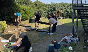 Several people work together in an outdoor garden space on a sunny day, assembling furniture and planting flowers. In the foreground, a person kneels on the grass filling a planter with soil beside large plant pots, while others assemble a black bench or shelving unit on a paved path. Gardening supplies, potted flowers, tools, and containers are arranged around the area. The space is bordered by mature shrubs and trees, with a metal staircase visible on the right, showing a community landscaping and improve