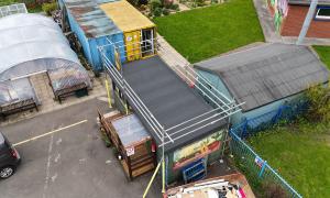 Aerial view of a community garden featuring a polytunnel greenhouse, storage buildings, shipping containers, and a small garden hub building surrounded by scaffolding