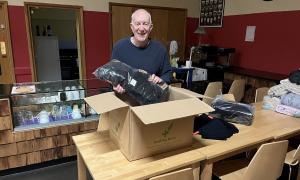Person unpacking donated clothing from a box in a community café, with additional garments laid out on tables and catering facilities in the background