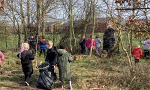 Children and adults participating in a woodland litter-picking and nature activity, carrying rubbish bags through a wooded area next to a grass field