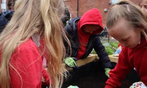 Several school-aged children wearing school uniforms and gardening gloves work together around a raised planting bed outdoors.