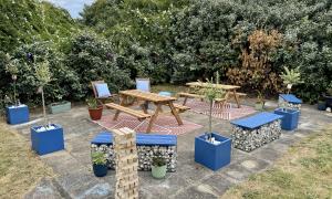 An outdoor community garden seating area on a paved patio, surrounded by dense green shrubs and climbing plants. Two wooden picnic benches sit on patterned outdoor rugs, with additional blue benches, planters containing small trees and flowers, and decorative gabion-style seating filled with stones. A stack of oversized wooden blocks stands in the foreground, and potted plants are placed throughout the space. The area appears designed for social gatherings and outdoor activities in a landscaped garden setti