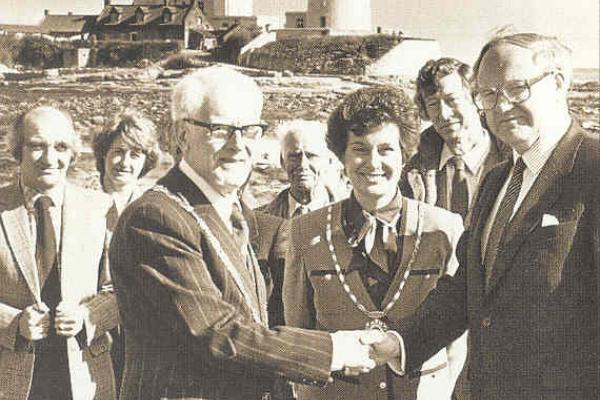 Two men shake hands infront of a crowd, a lighthouse is in the background