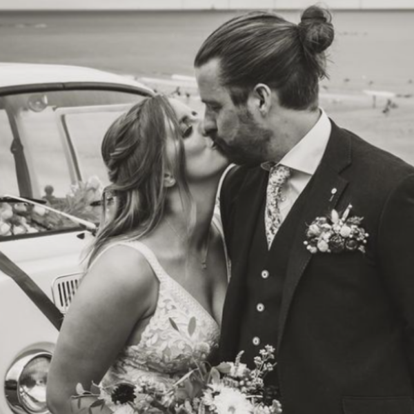 A black and white photo of a newly wed couple kissing infront of a white car. 
