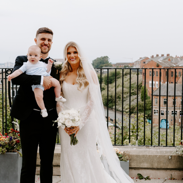 A bride and groom standing on a balcony. The groom is holding a baby